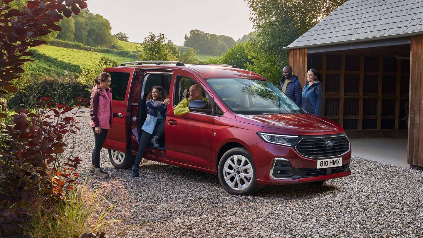 Ford Grand Tourneo Connect rosso, vista frontale a tre quarti, parcheggiato in un vialetto ghiaioso davanti a una casa