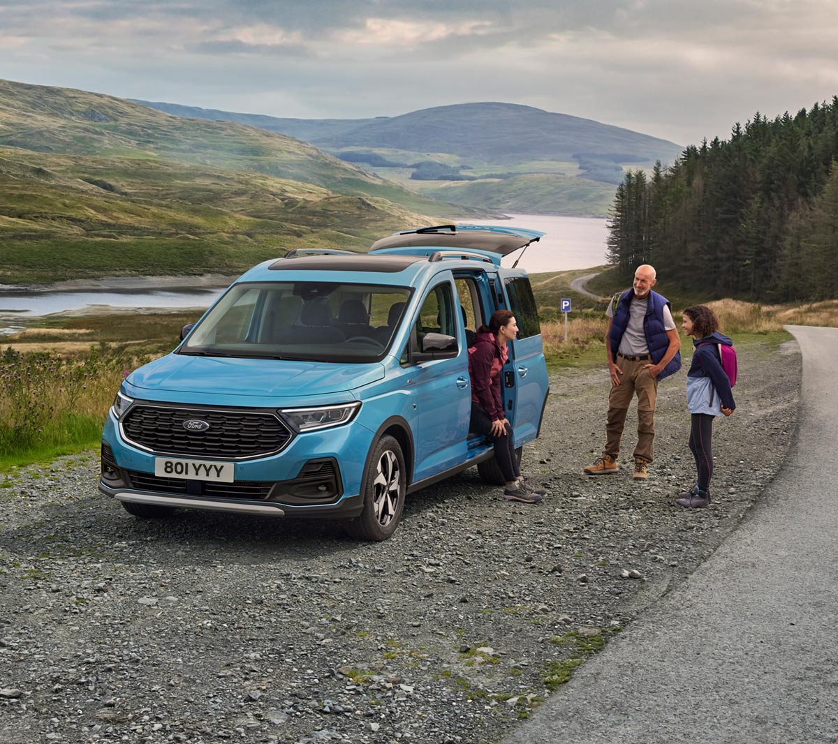 Ford Tourneo Connect Active  azzurro, vista frontale a tre quarti con porta scorrevole aperta, parcheggiato su una strada sterrata. Una donna è seduta a bordo, fuori vicino a lei un uomo e una ragazza