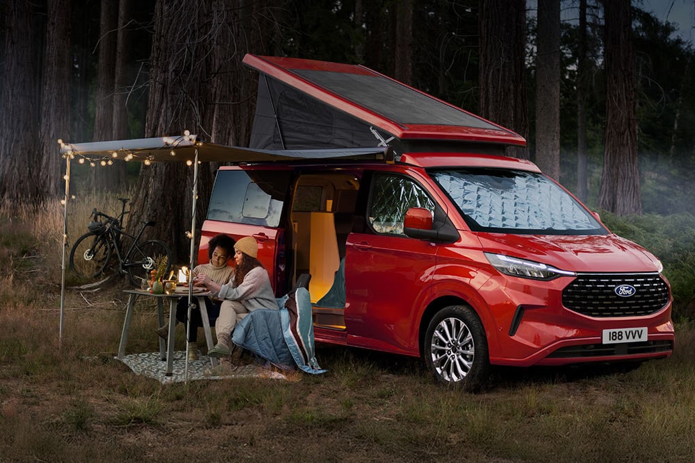 Due donne sedute sotto una tenda davanti alle portiere laterali aperte di un Ford Nugget con tetto inclinabile aperto.