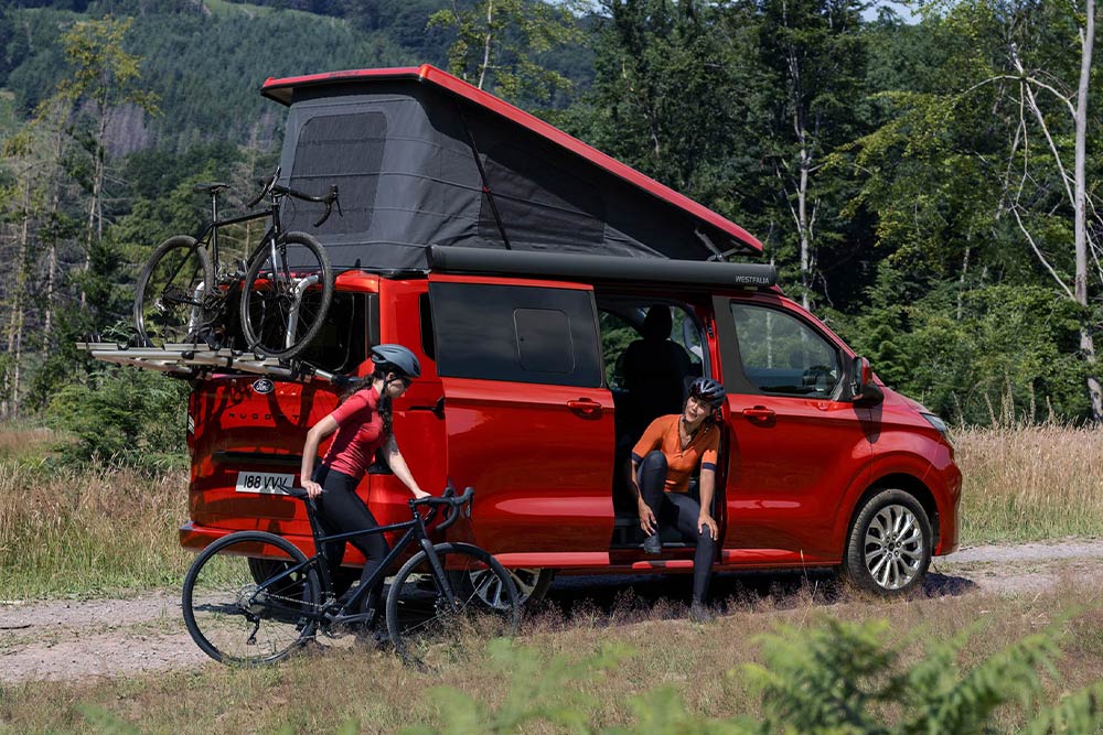 Alcune persone scaricano le biciclette dal portabici di un Ford Nugget rosso.