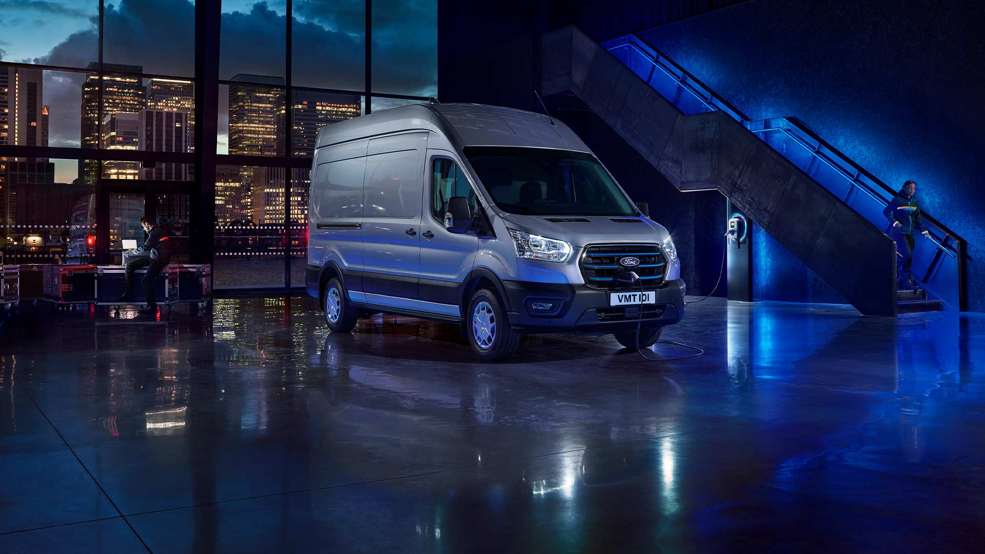 Il furgone elettrico Ford E-Transit color argento è parcheggiato al chiuso con un profilo della città visibile attraverso grandi finestre. 