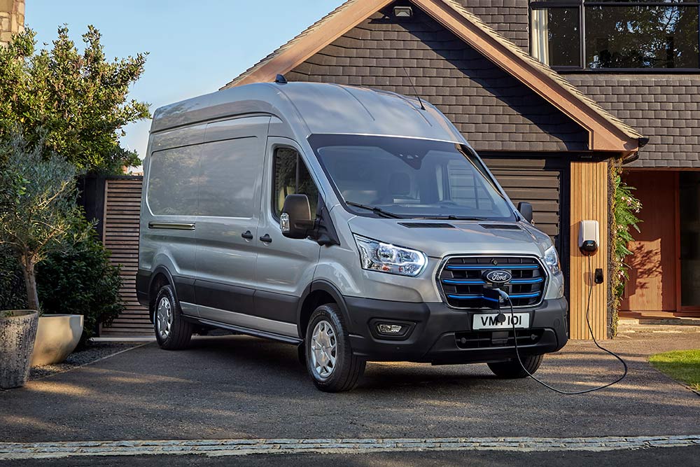 Il furgone Ford E-Transit di color argento è parcheggiato in un vialetto accanto a una casa, collegato a una stazione di ricarica.