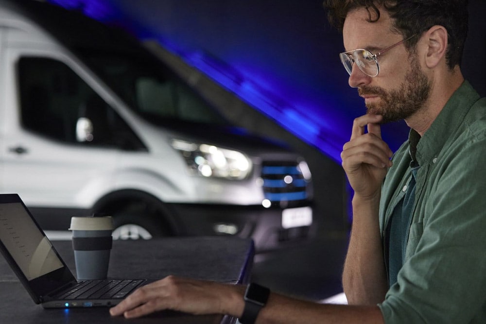 Uomo che lavora su un laptop con un furgone Ford E-Transit sullo sfondo.