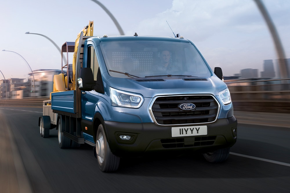 Vista della griglia anteriore di un Ford Transit Chassis Cab che trasporta un escavatore lungo una strada cittadina. 