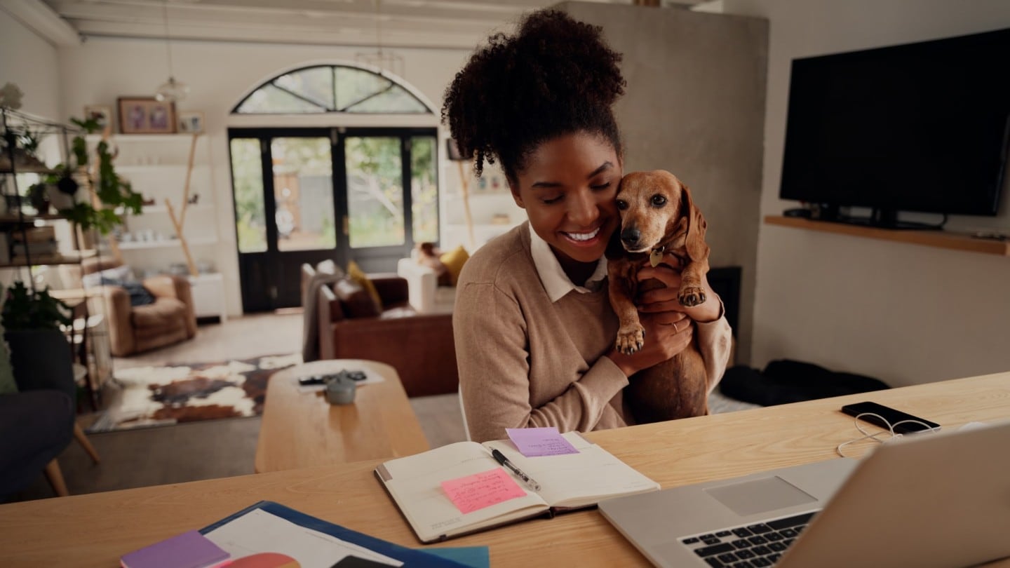 Donna in casa con cane in braccio che sorride davanti a un computer portatile