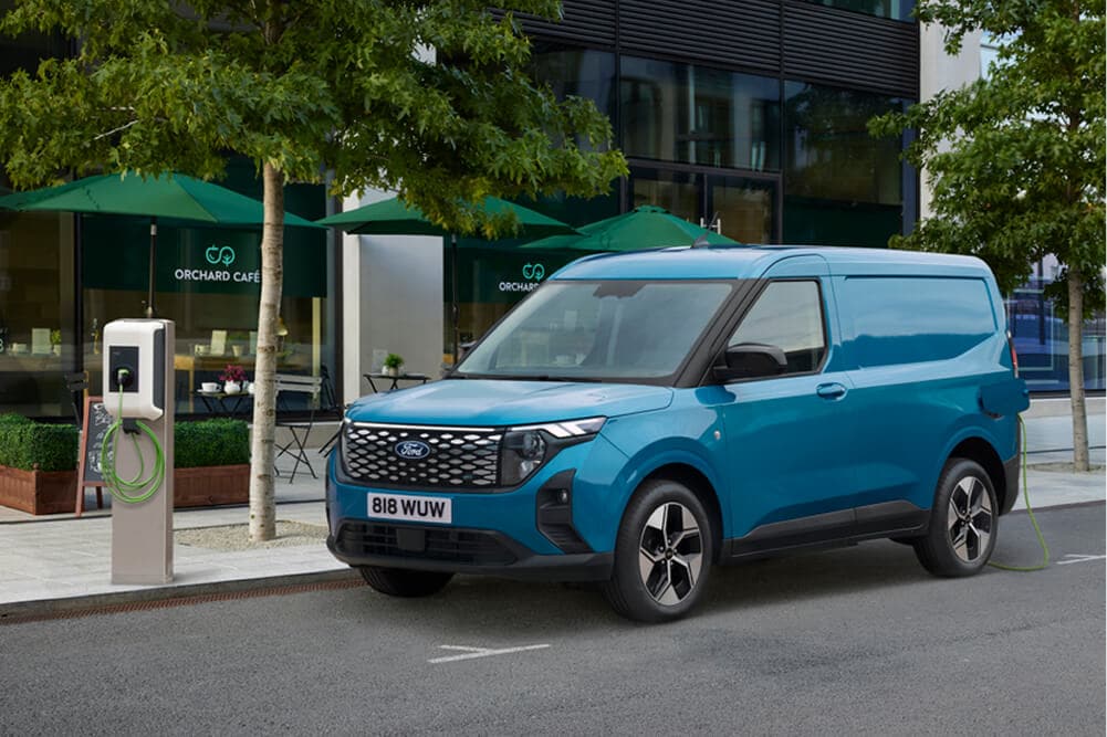 Ford E-Transit Courier blu, vista anteriore a tre quarti, parcheggiato accanto a una colonnina di ricarica