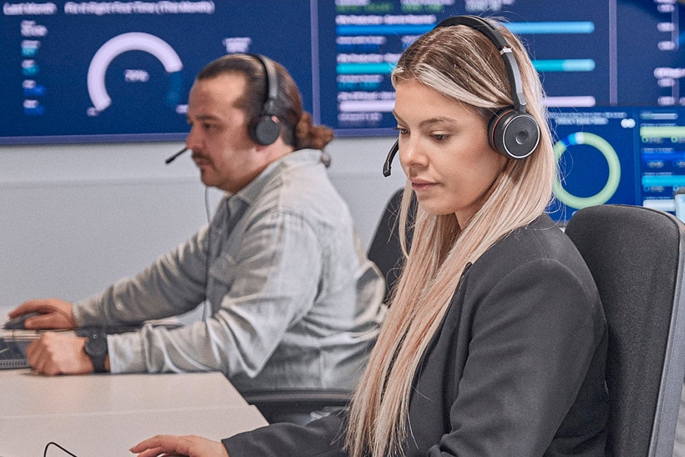 Due persone con cuffie lavorano al computer in un ufficio con schermi di dati sullo sfondo.