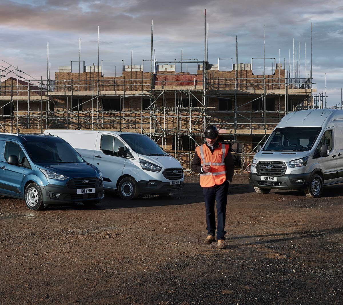 Lavoratore con casco da cantiere e gilet ad alta visibilità davanti a un cantiere con veicoli Ford sullo sfondo