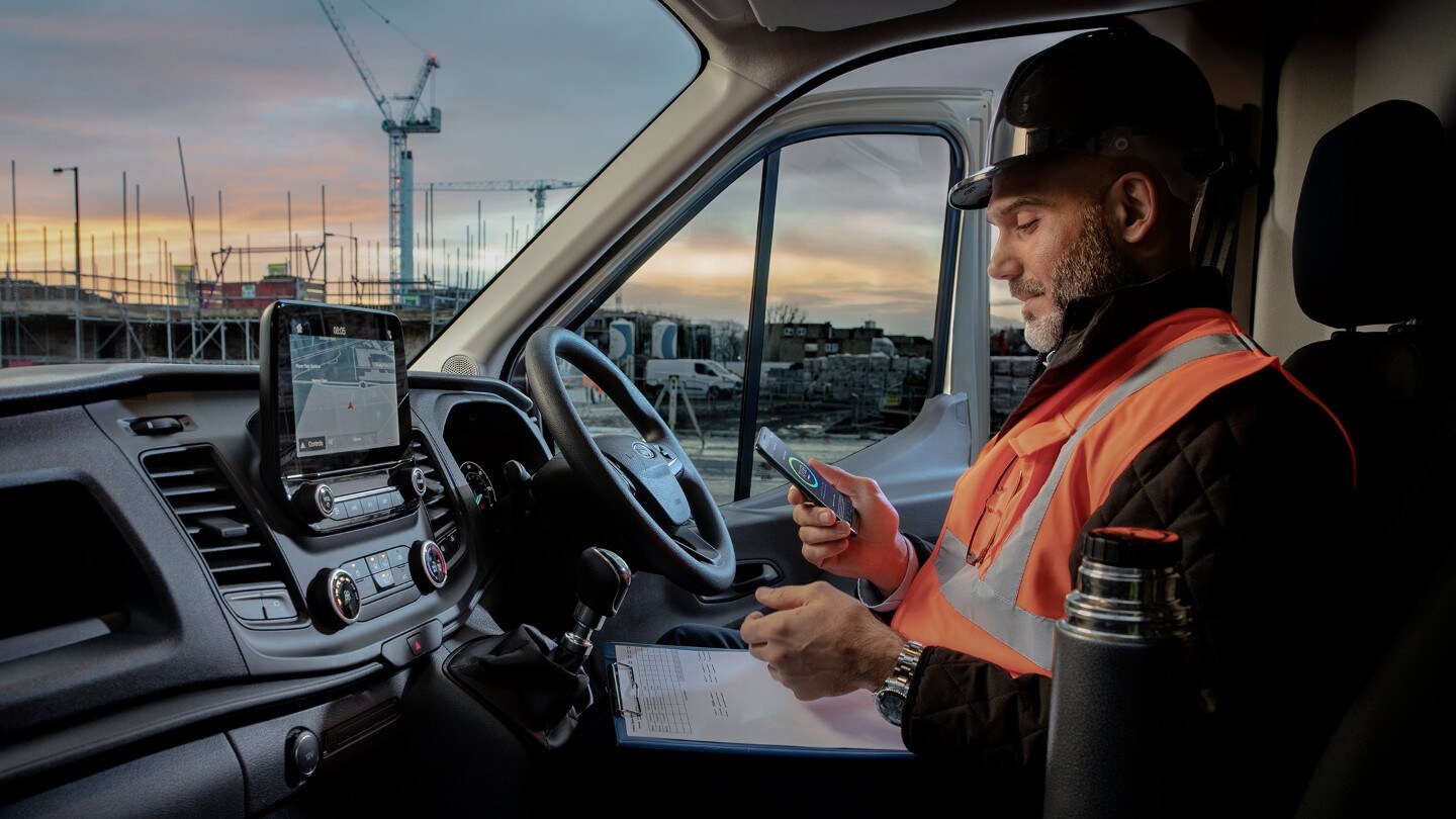 Operaio con casco da cantiere siede al volante di un veicolo guardando un telefono cellulare 