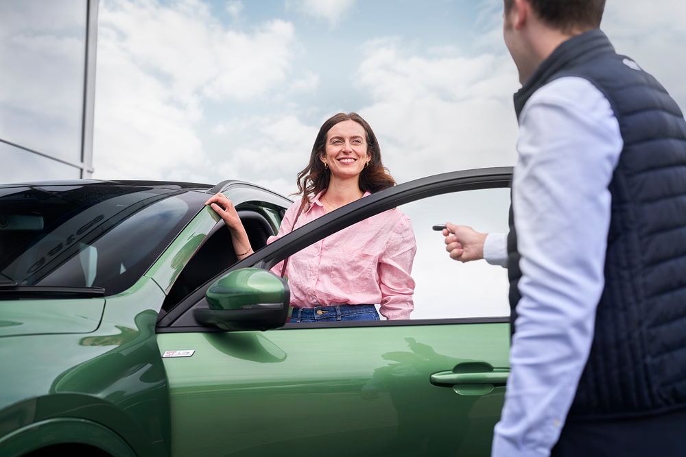 Una donna con una camicia rosa sorride accanto a un'auto verde, con un concessionario Ford che le consegna le chiavi dell'auto.