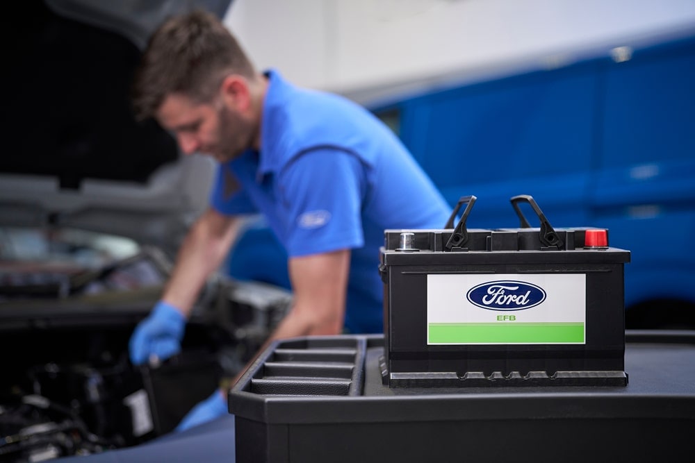 Un meccanico Ford al lavoro sul motore di un'auto, con una batteria Ford in primo piano.