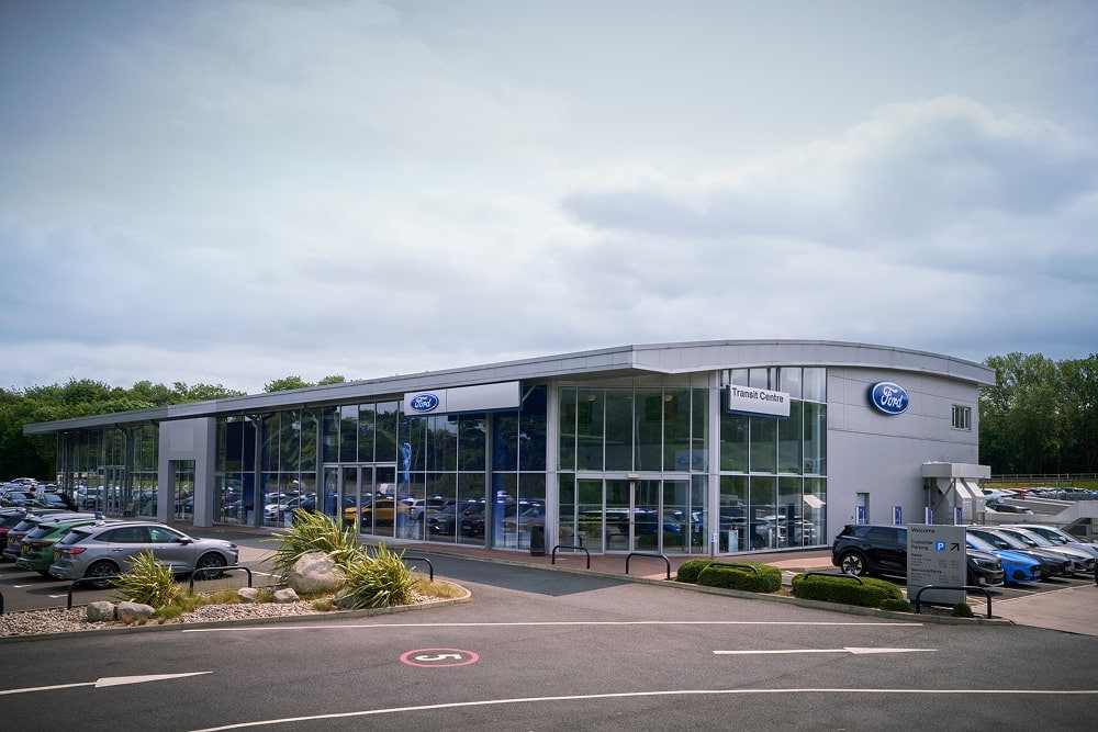 Vista dall'esterno di una concessionaria Ford con grandi vetrate. Le auto sono parcheggiate all'esterno.