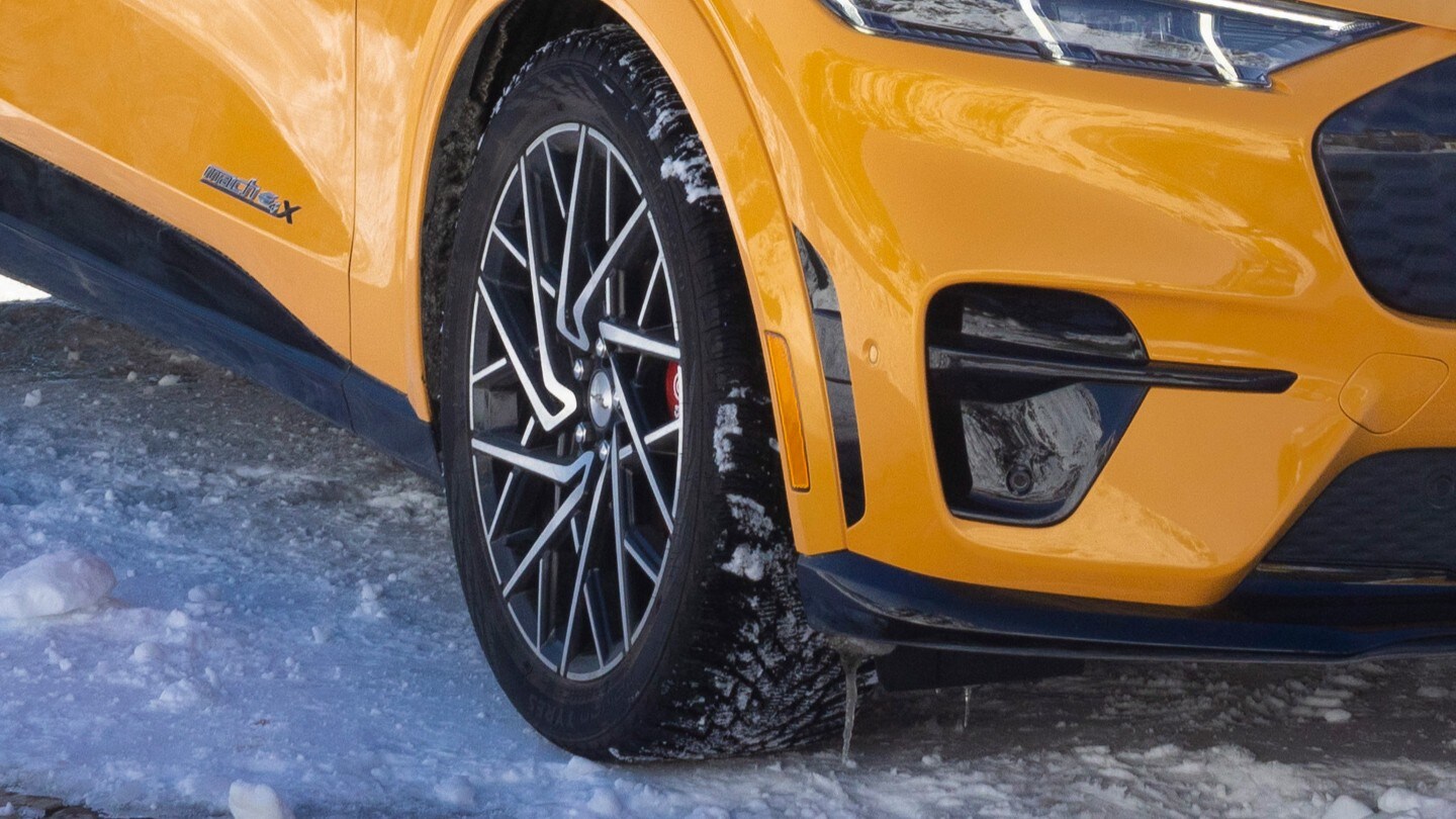 Primo piano della ruota frontale e del parafango di un’auto color giallo vivace, come una Ford Mustang Mach-E, parcheggiata su un manto stradale innevato.